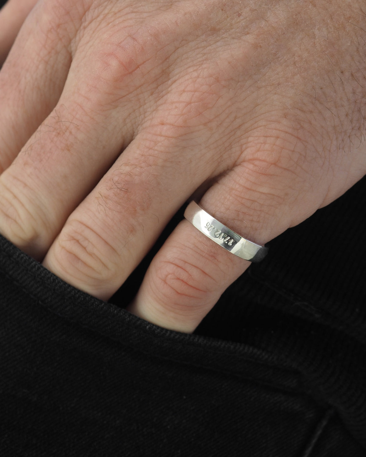 A close-up of a man's hand wearing a polished silver band ring engraved with the numbers.