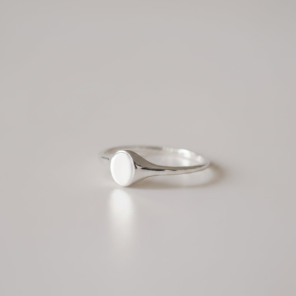 Gold signet ring with a smooth, round face on a sleek band, set against a light grey background. Simple and elegant jewellery design.