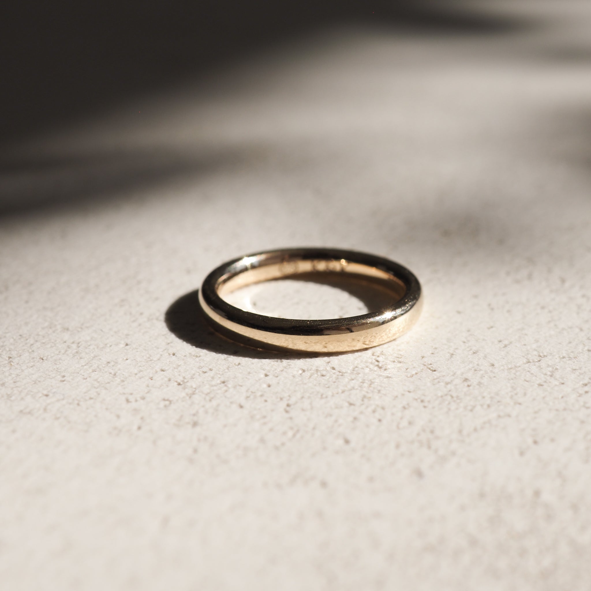 Elegant gold ring on textured beige surface, casting a soft shadow in natural light. Simple yet luxurious jewellery piece.