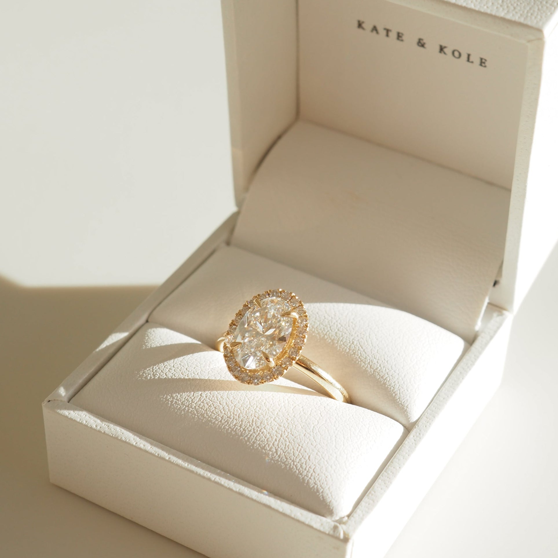 A gold engagement ring featuring an oval-shaped centre diamond surrounded by a delicate halo of smaller diamonds, displayed in a white leather Kate & Kole ring box with soft natural lighting.