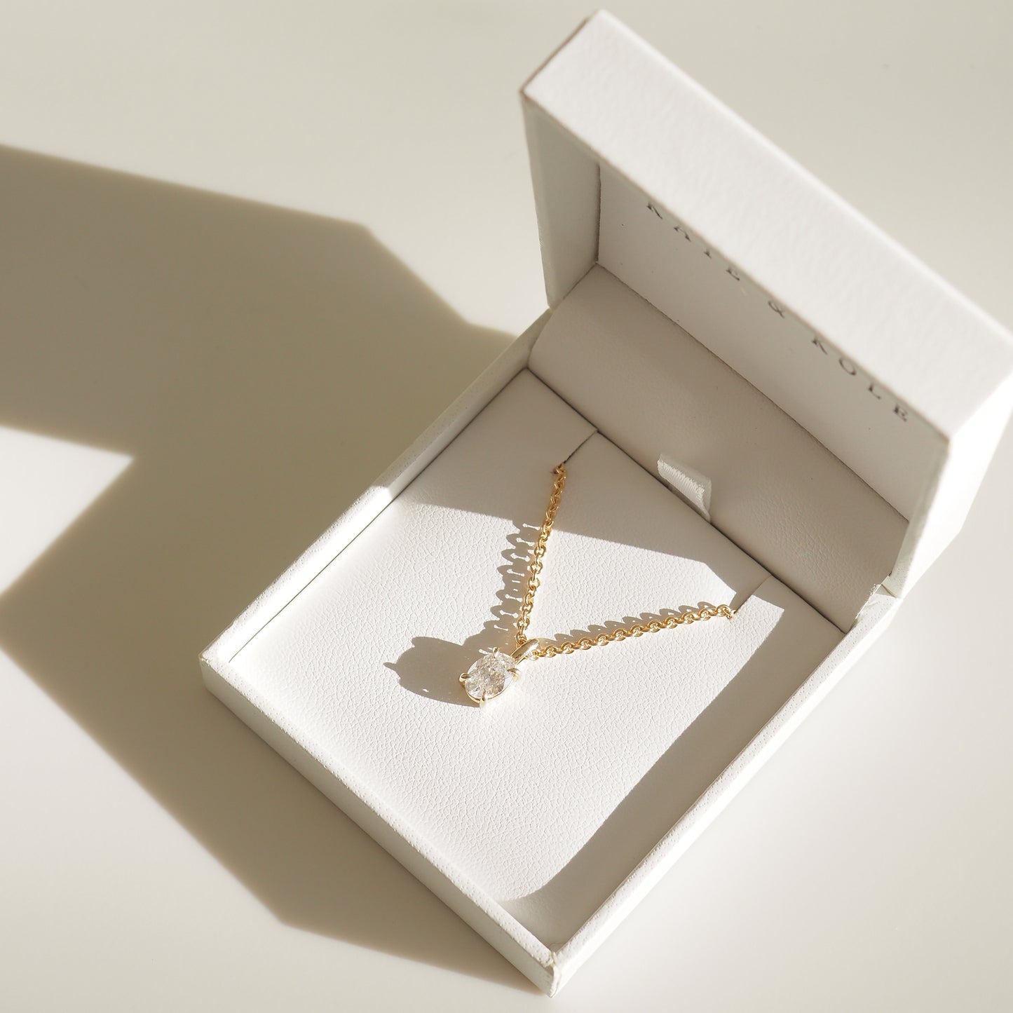 Gold chain necklace with a diamond pendant in an elegant white jewellery box, casting a soft shadow on a light surface.
