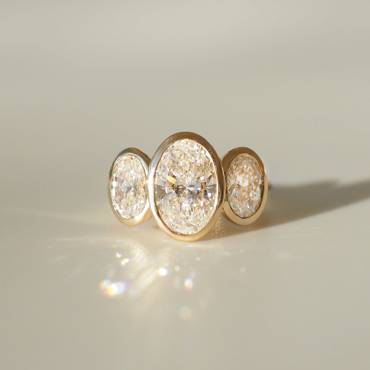 Gold ring with three oval-cut diamonds set in bezel settings, elegantly reflecting light against a soft beige background.