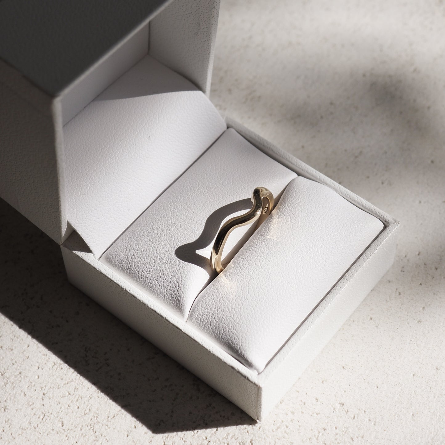 A gold wave-shaped ring displayed in an open white jewellery box on textured surface, highlighting luxury design and craftsmanship.