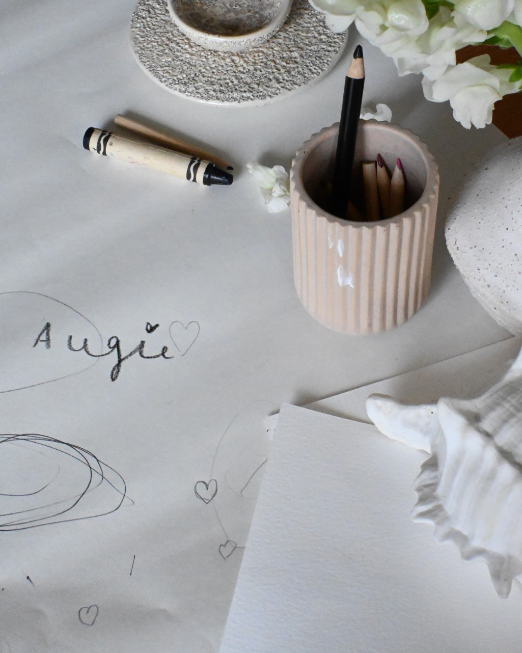 Sketch pad with "Augie" and heart doodles, surrounded by pencils in a ceramic holder, crayons, white flowers, and textured decor.