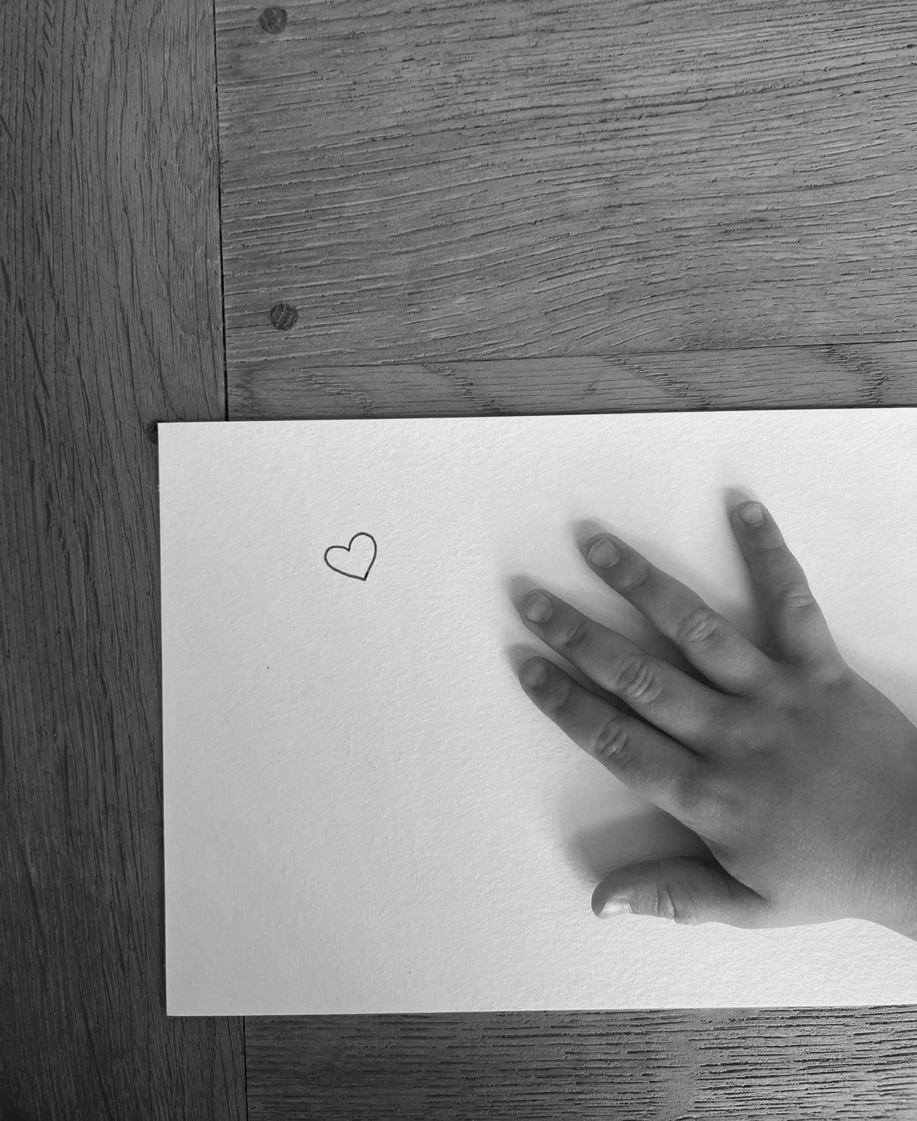 Child's hand resting on white paper with a small heart sketch, placed on a wooden surface. Black and white photo.