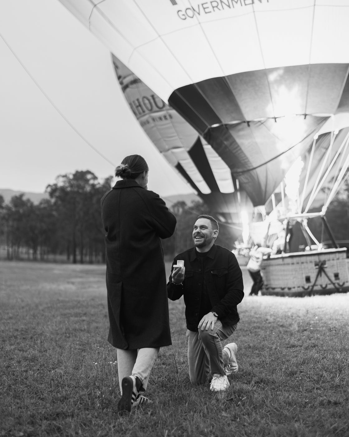 A man kneels, proposing with a jewellery box to a woman in front of an inflated hot air balloon in a scenic field. Black and white image.