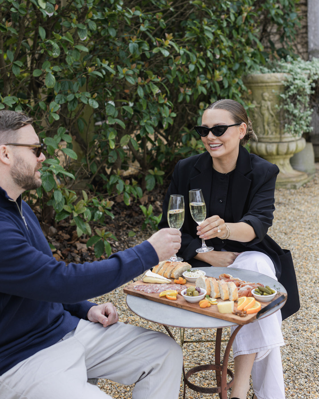 Two people enjoying champagne outdoors at a stylish cafe table, adorned with a gourmet charcuterie board. Elegant, luxury jewellery scene.
