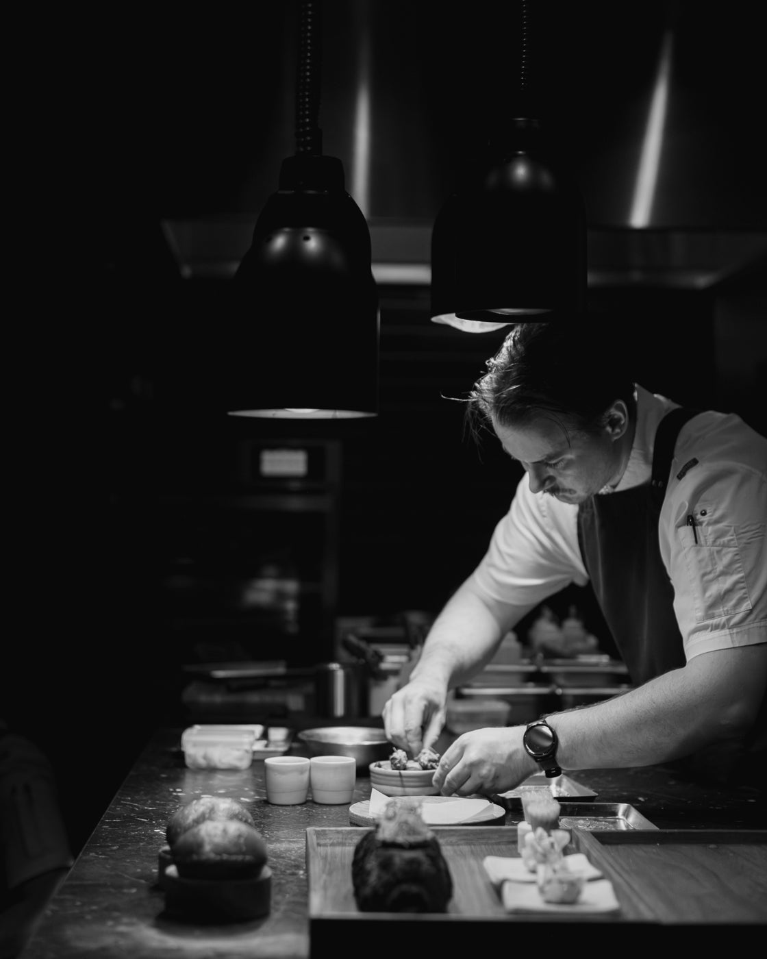 Chef crafting gourmet dish in dimly lit kitchen. Focused on fine plating, surrounded by cooking elements. Culinary artistry emphasised.