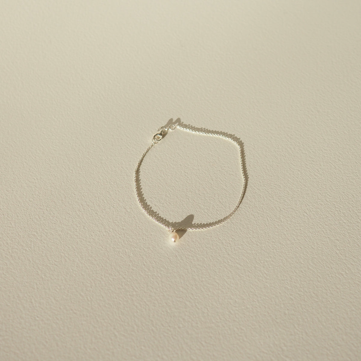 Delicate gold chain bracelet with a single pearl pendant, casting a soft shadow on a light-coloured surface. Minimalist luxury jewellery.