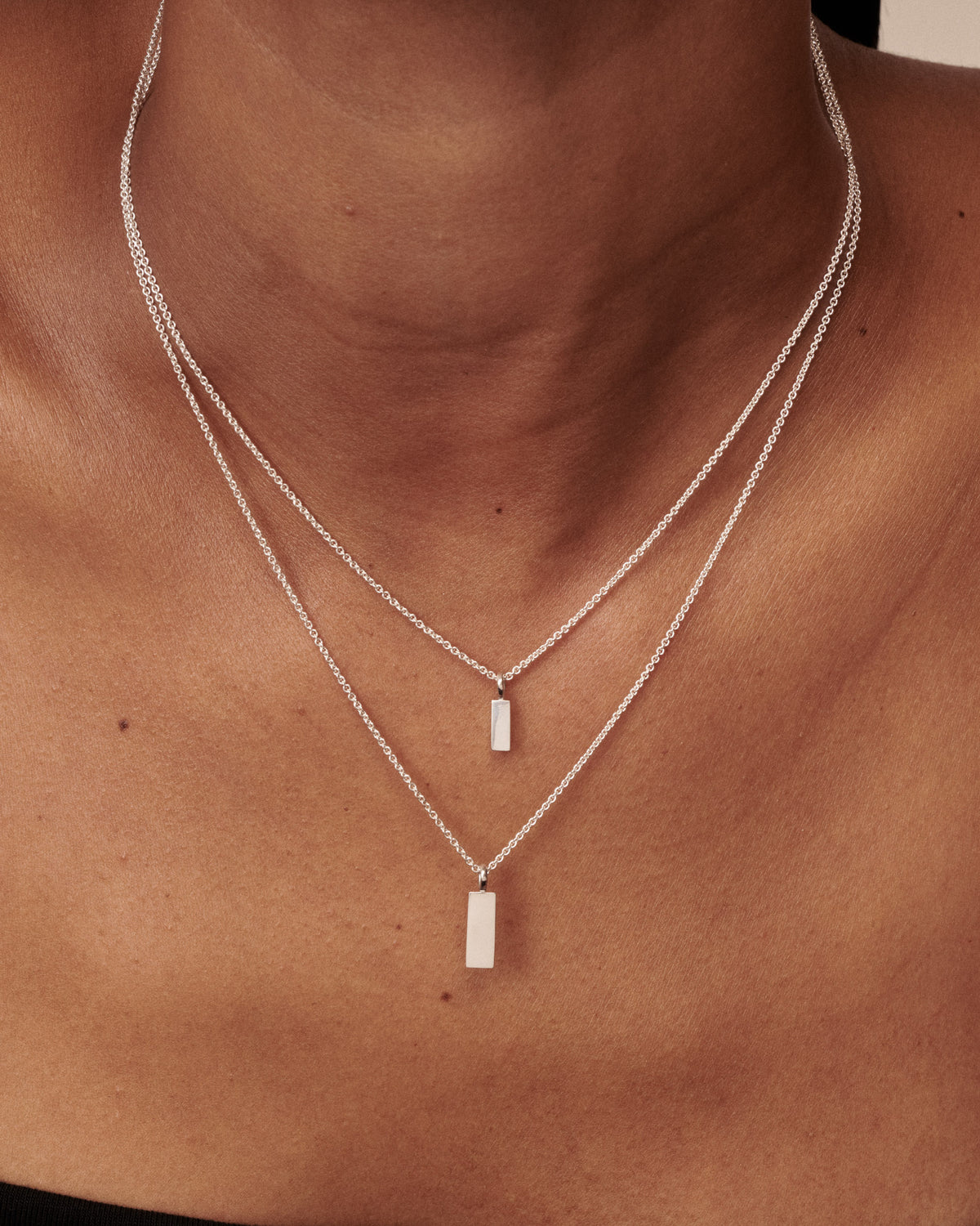 A close-up of a person's neck and upper chest wearing a delicate double-layered silver necklace with rectangular bar pendants. [text overlay: Small (top) Pendant & Large (bottom) Pendant]
