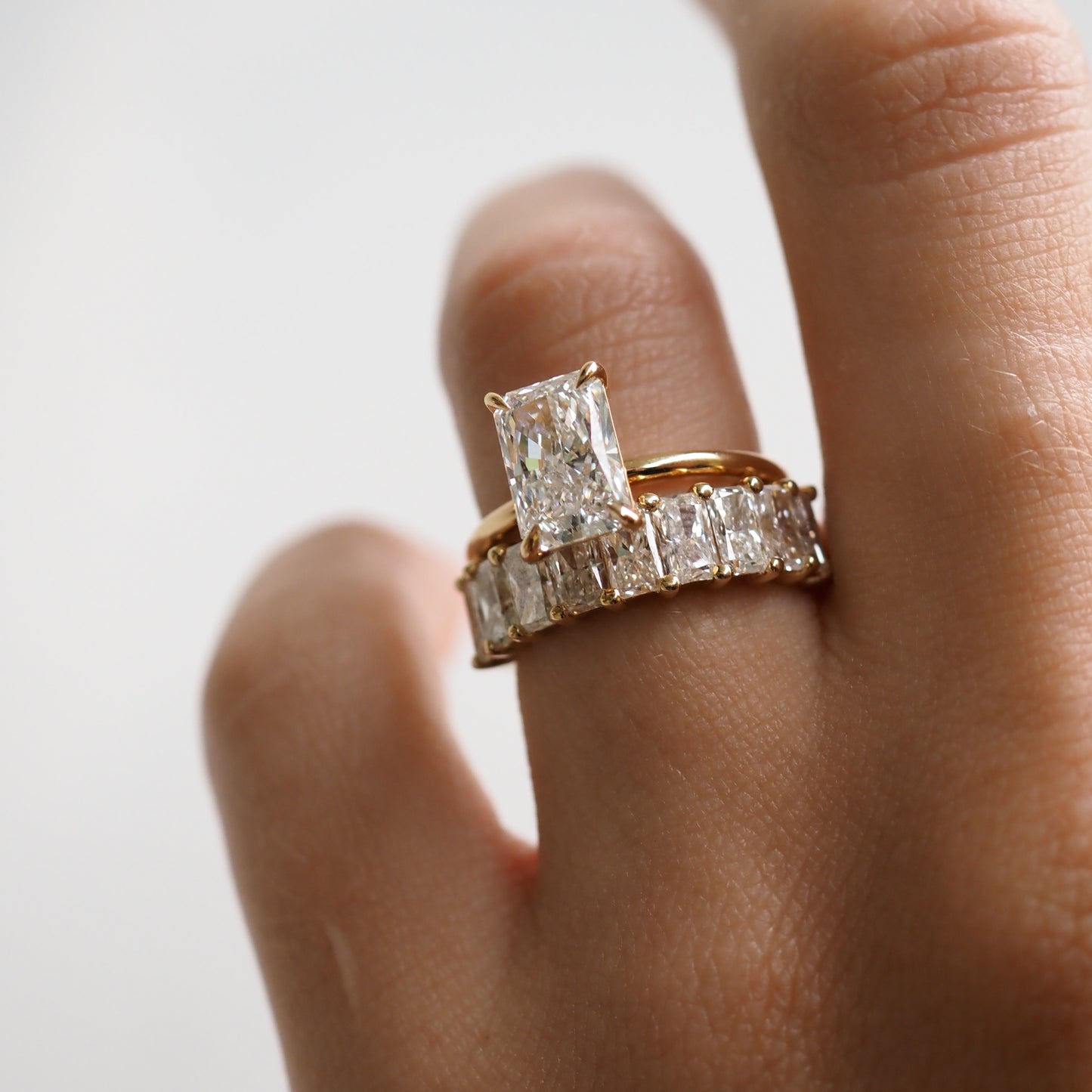 Elegant hand adorned with a gold band featuring a large rectangular diamond and a matching eternity ring, exemplifying luxury jewellery design. [text overlay: 1.5ct Khai and Radiant Lace] [text colour: #000000]
