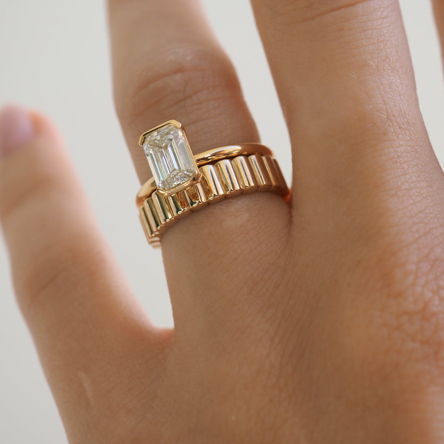 Gold rings with an emerald-cut clear stone on a person's ring finger. The rings have a ribbed design, showcasing luxury jewellery.