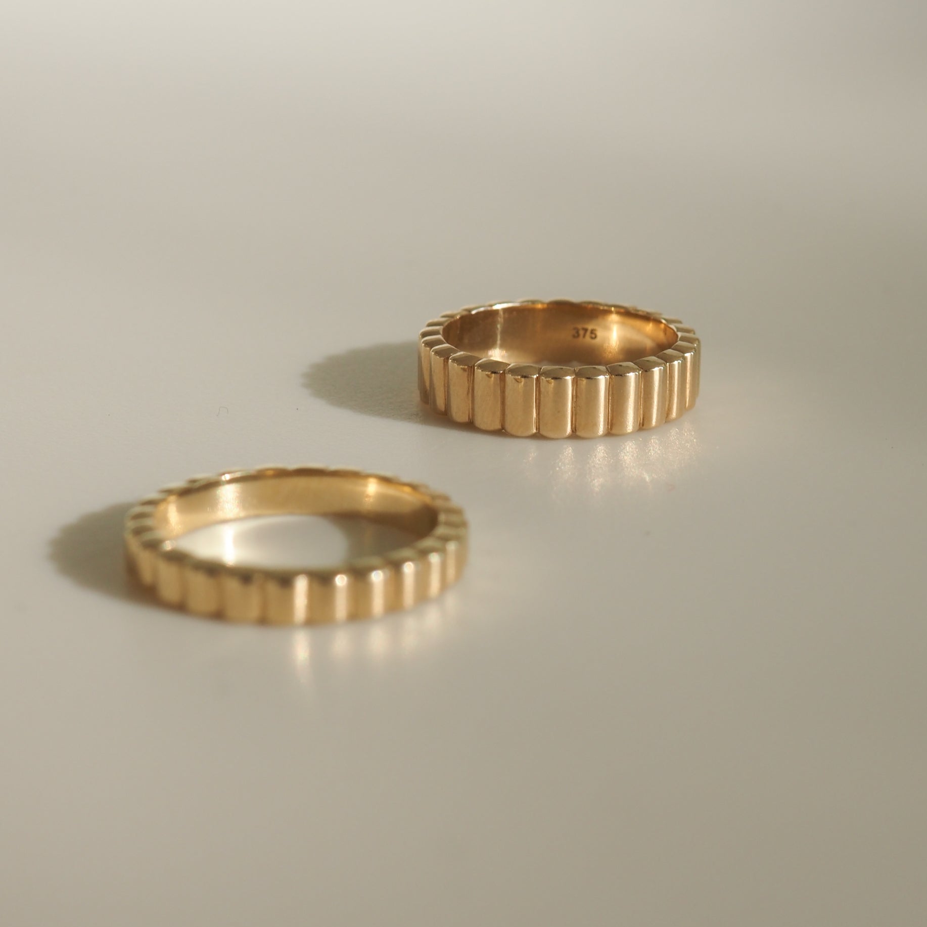 Two ribbed gold rings on a light surface, one standing upright showing "375" hallmark inside. Luxurious jewellery, minimalistic design.