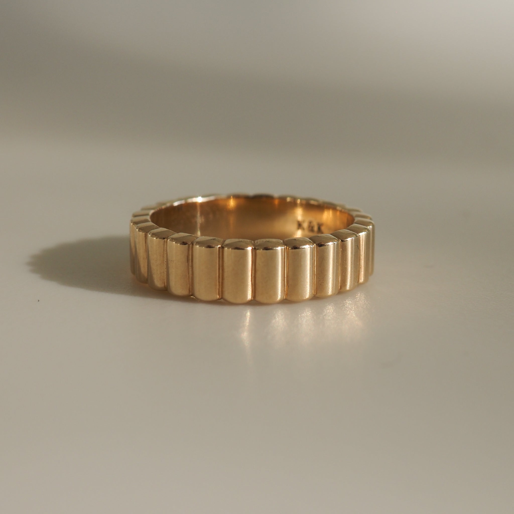 Elegant gold ring with a ribbed design, reflecting light on a plain surface. Perfect for luxury jewellery lovers.