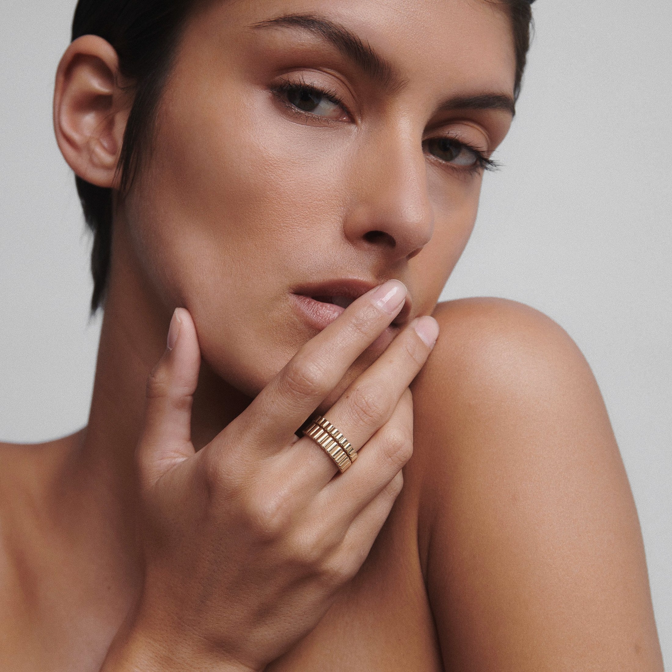Model with short hair showcases a stack of luxury gold rings on her fingers against a plain background, highlighting elegant jewellery design.