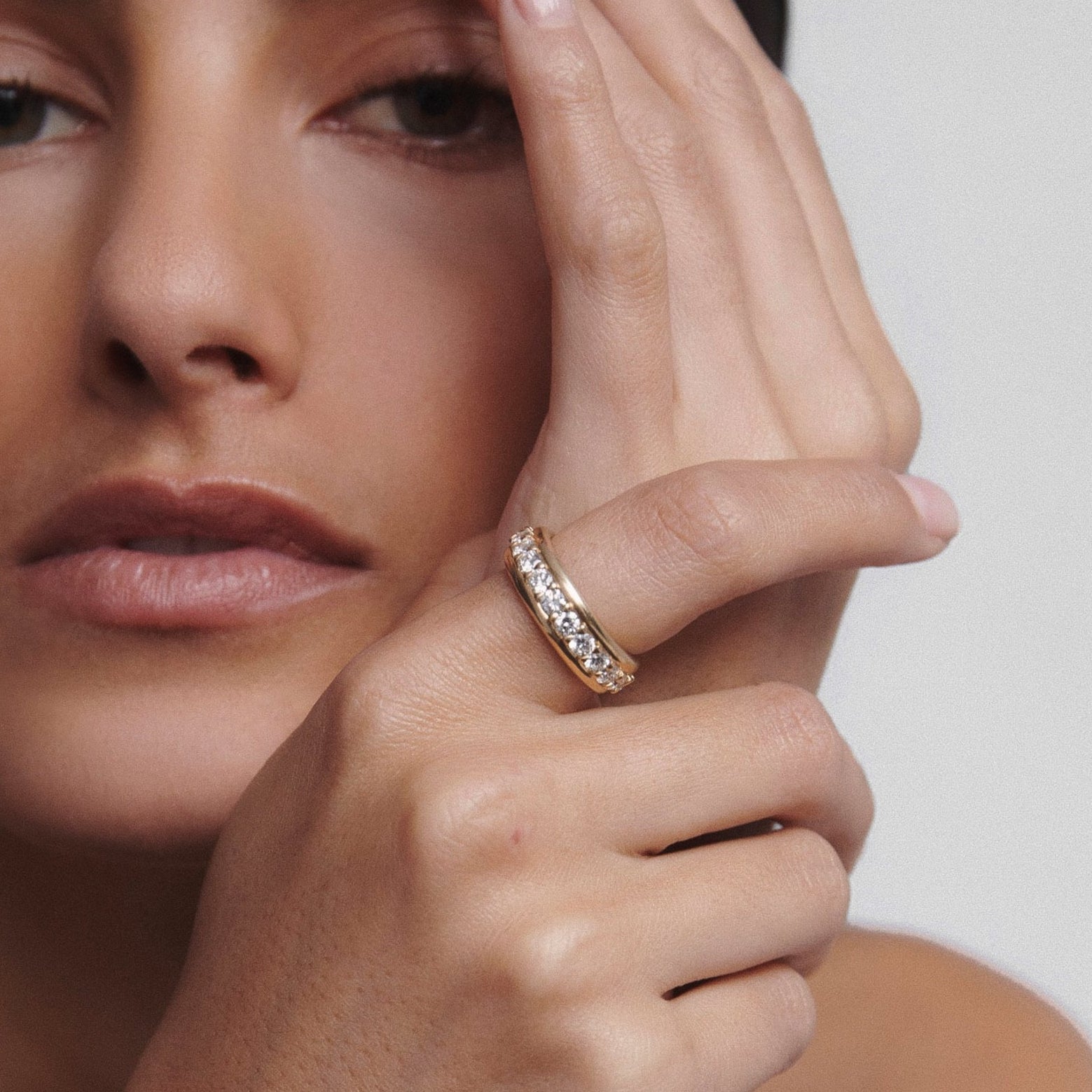 Woman with minimal makeup showcasing a gold diamond ring on her finger, hand resting on her face, set against a plain background.