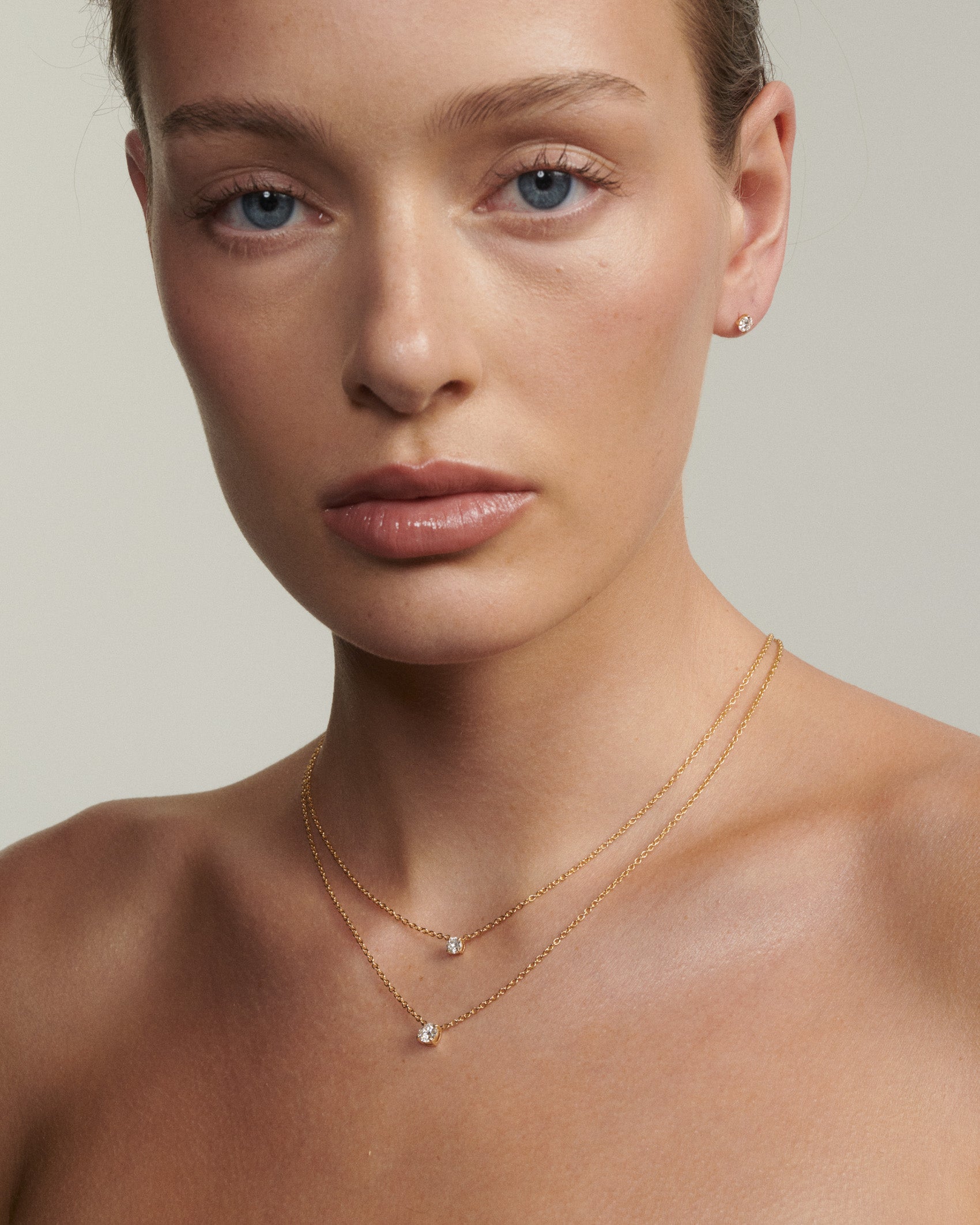 Close-up of a woman wearing a delicate gold necklace on a neutral background [text overlay: Andrea wears the 0.25ct & 0.5ct Round Diamond Necklace]