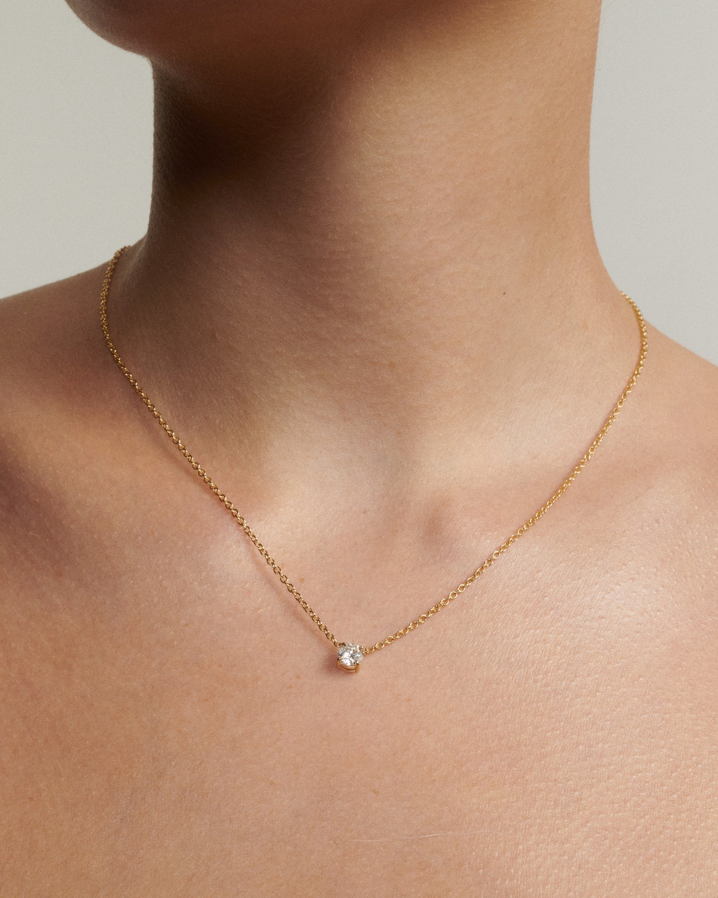 Gold necklace with a small gemstone on a person's neck against a neutral background [text overlay: Andrea wears the 0.5ct Round Diamond Necklace]