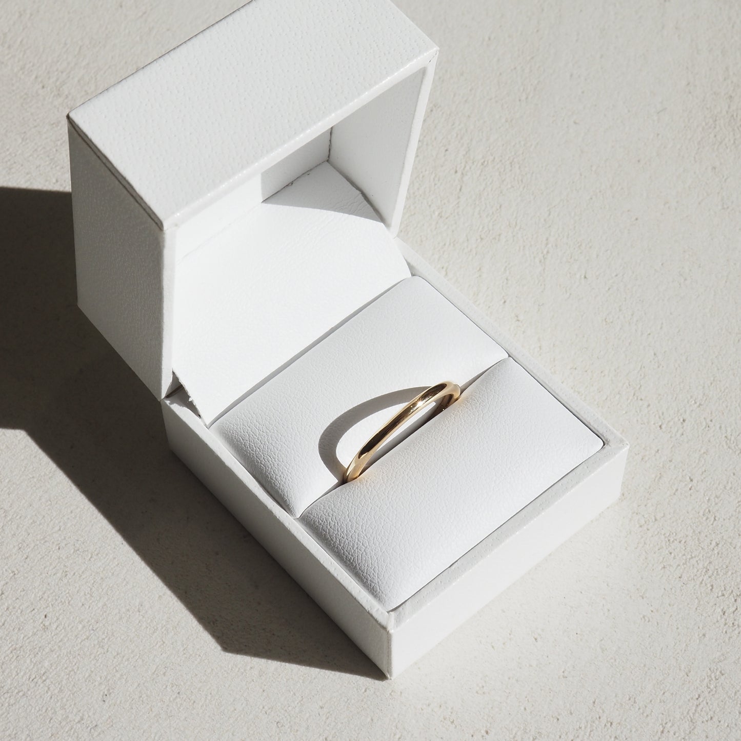 Elegant gold ring in a white leather jewellery box, sunlight casting a shadow, emphasising its luxury and minimalist design.