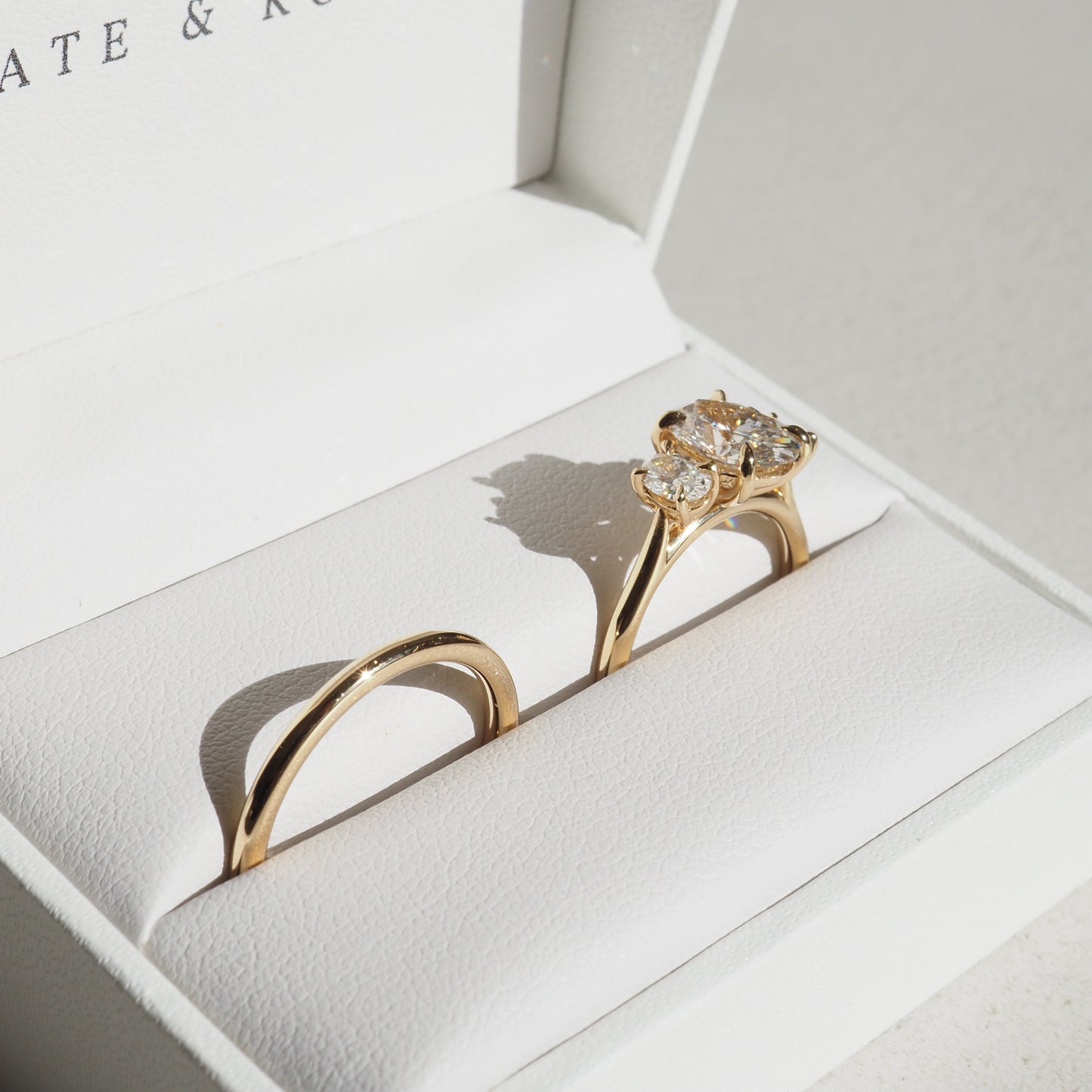 Elegant gold engagement ring set with accent diamonds, displayed in a luxe white jewellery box, capturing sunlight and casting delicate shadows.