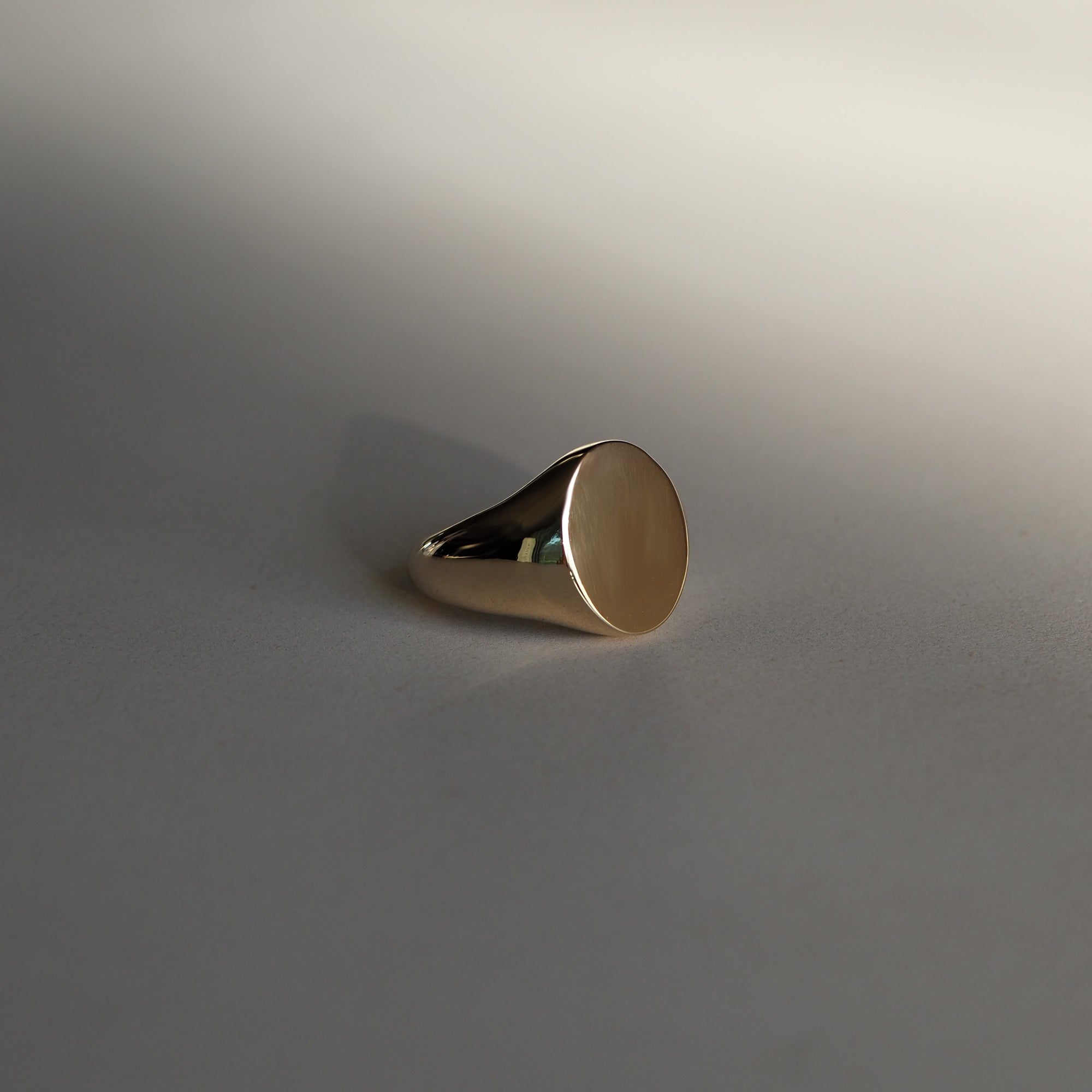 Elegant gold signet ring with a smooth, polished face on a soft grey background, showcasing high-end luxury jewellery design.