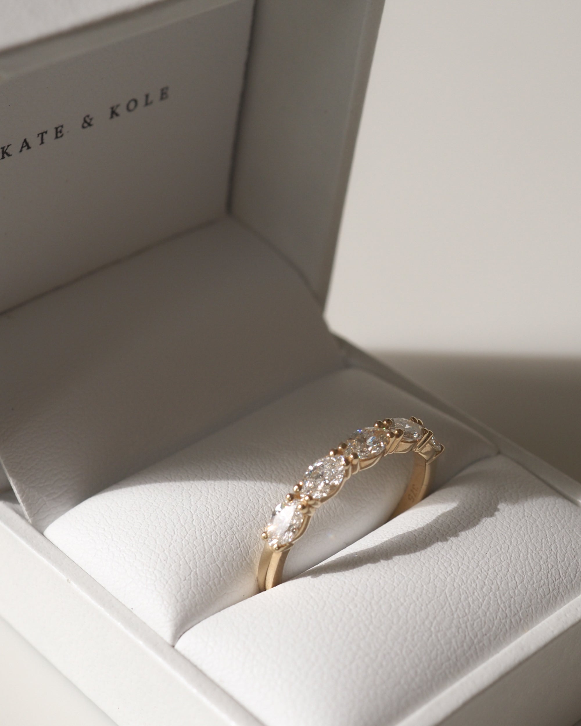 A gold ring with five marquise-cut diamonds in a white jewellery box, branded "Kate & Kole", under soft lighting.