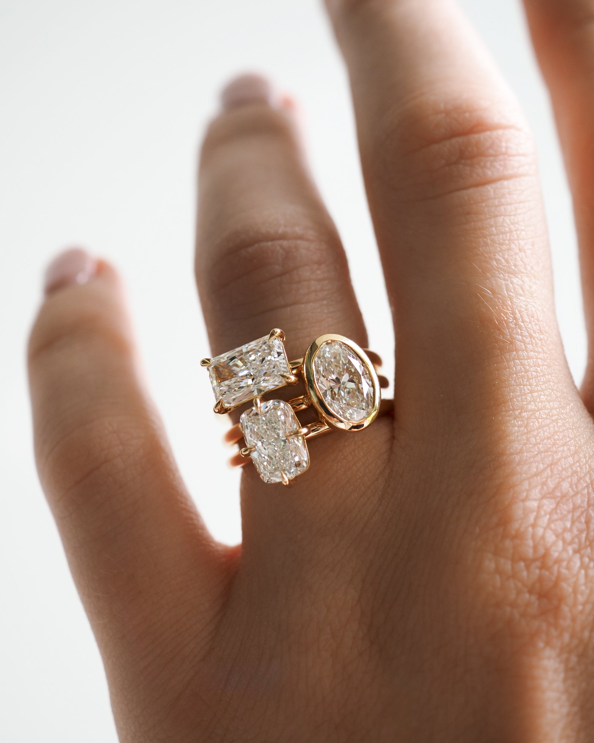 Close-up of a hand adorned with three luxury gold rings, each showcasing a unique diamond cut: rectangular, oval, and emerald.