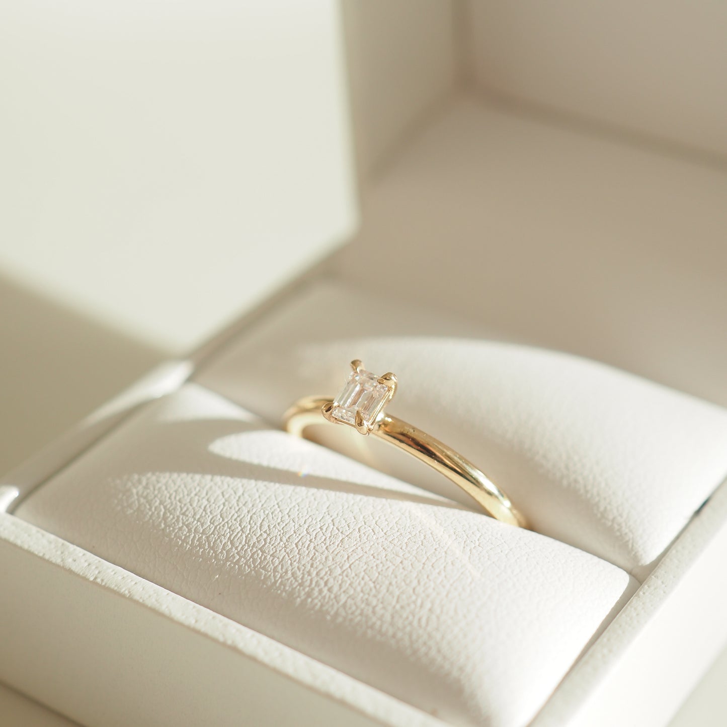 Gold ring with a rectangular diamond in an elegant white jewellery box, captured under soft sunlight. [text overlay: Emerlad cut lab-grown diamond]