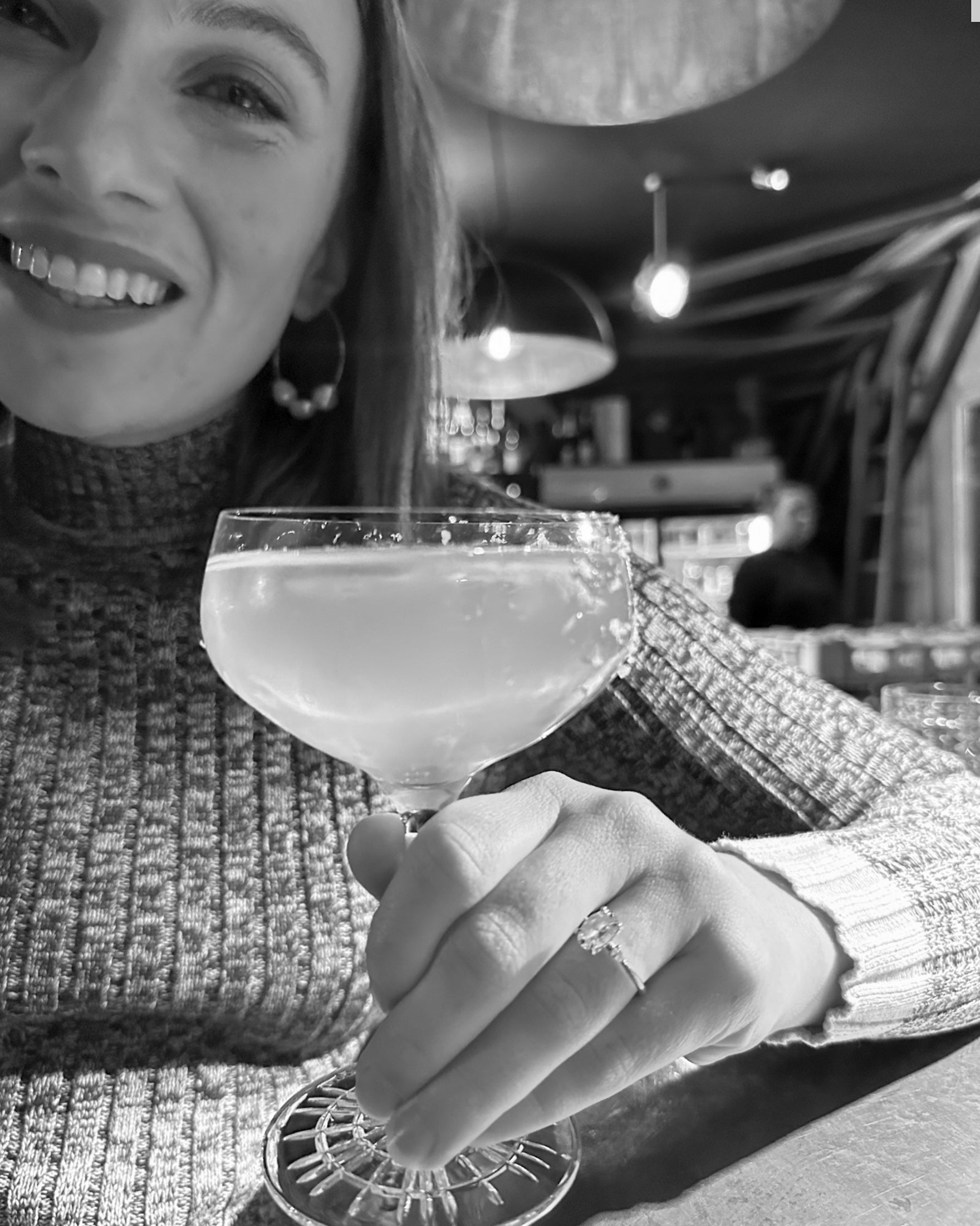 Woman in a cosy bar holds a cocktail, showcasing an elegant diamond ring. Soft lighting and a stylish knit jumper complete the luxe vibe.