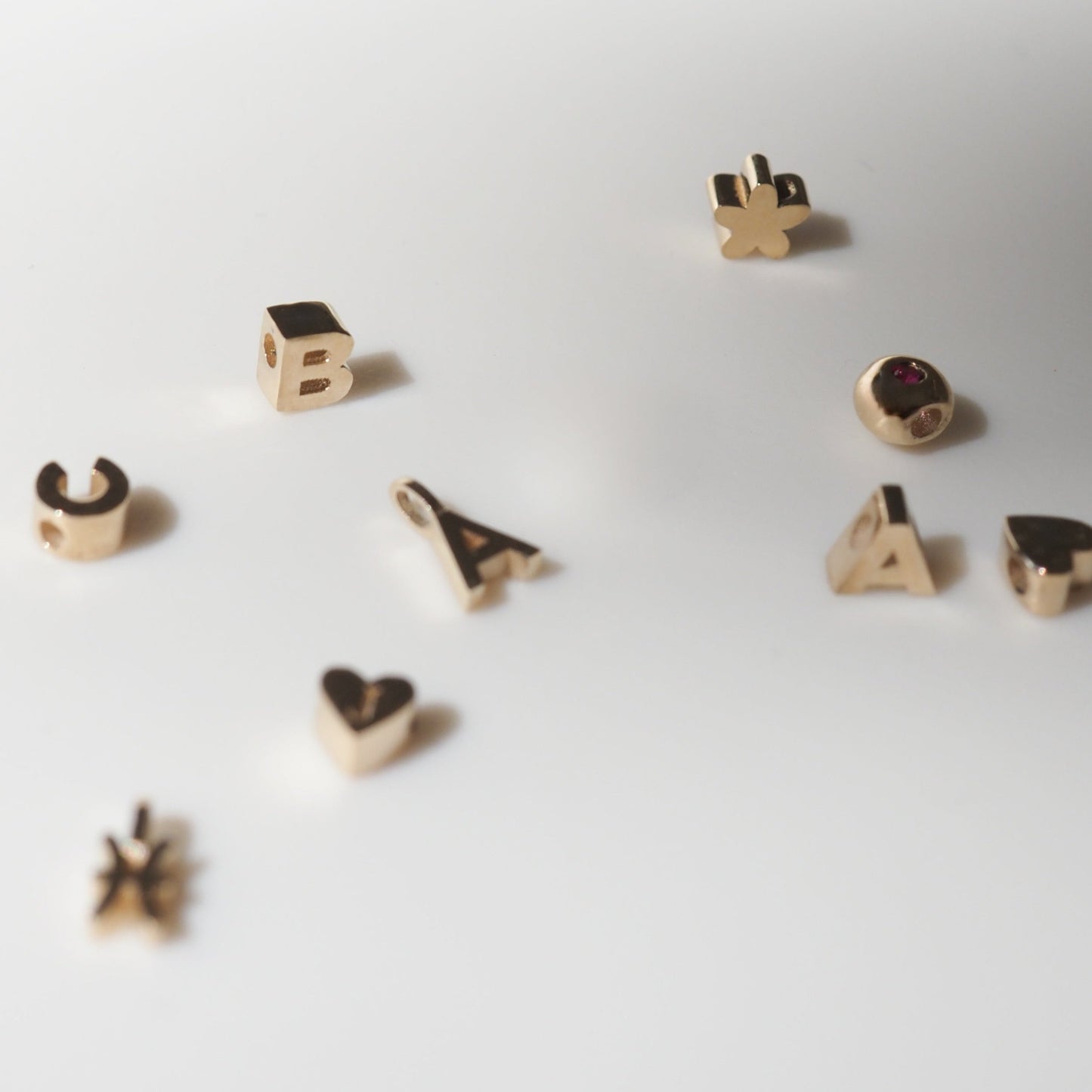 Gold pendant charms in various shapes including letters, a heart, and a star, scattered on a white surface. Luxurious jewellery elements.
