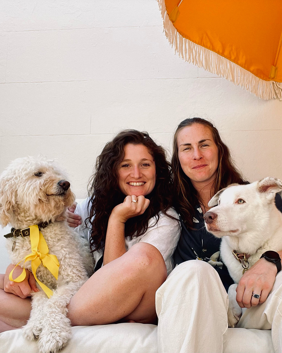 Two smiling women seated with two dogs under a vibrant orange umbrella. The dogs wear yellow and tan accessories, exuding a relaxed vibe.