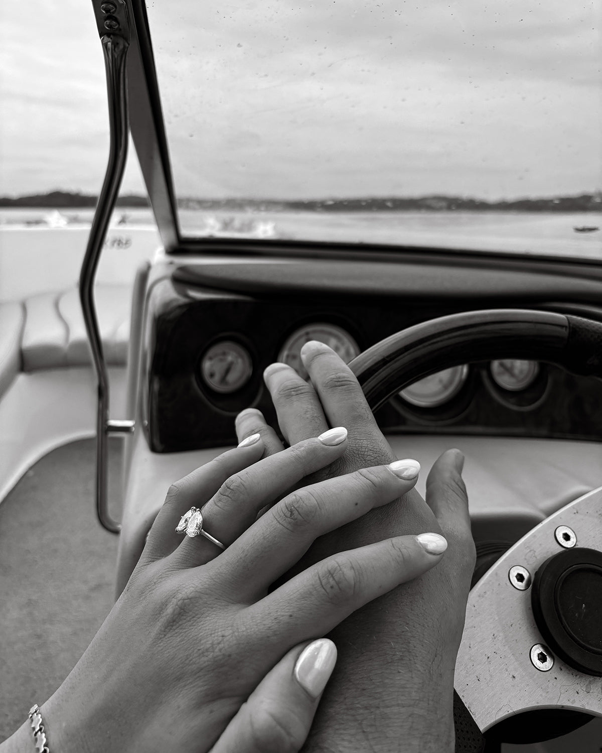 Couple holding hands on a boat steering wheel, showcasing a sparkling diamond engagement ring. Scenic water view in the background.