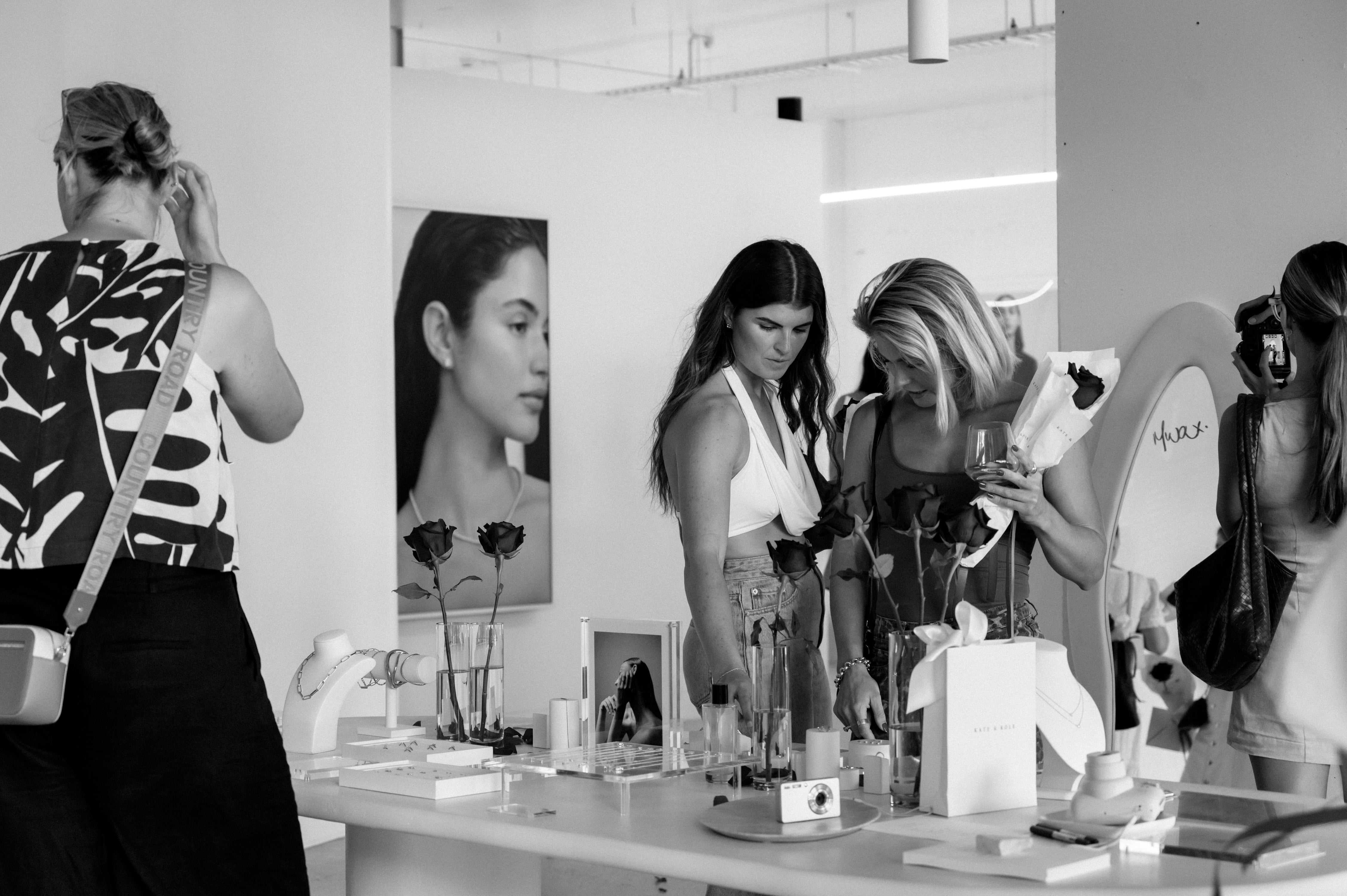 A monochrome photo of women examining luxury jewellery and cosmetics in a stylish boutique, with a portrait and roses in the background.