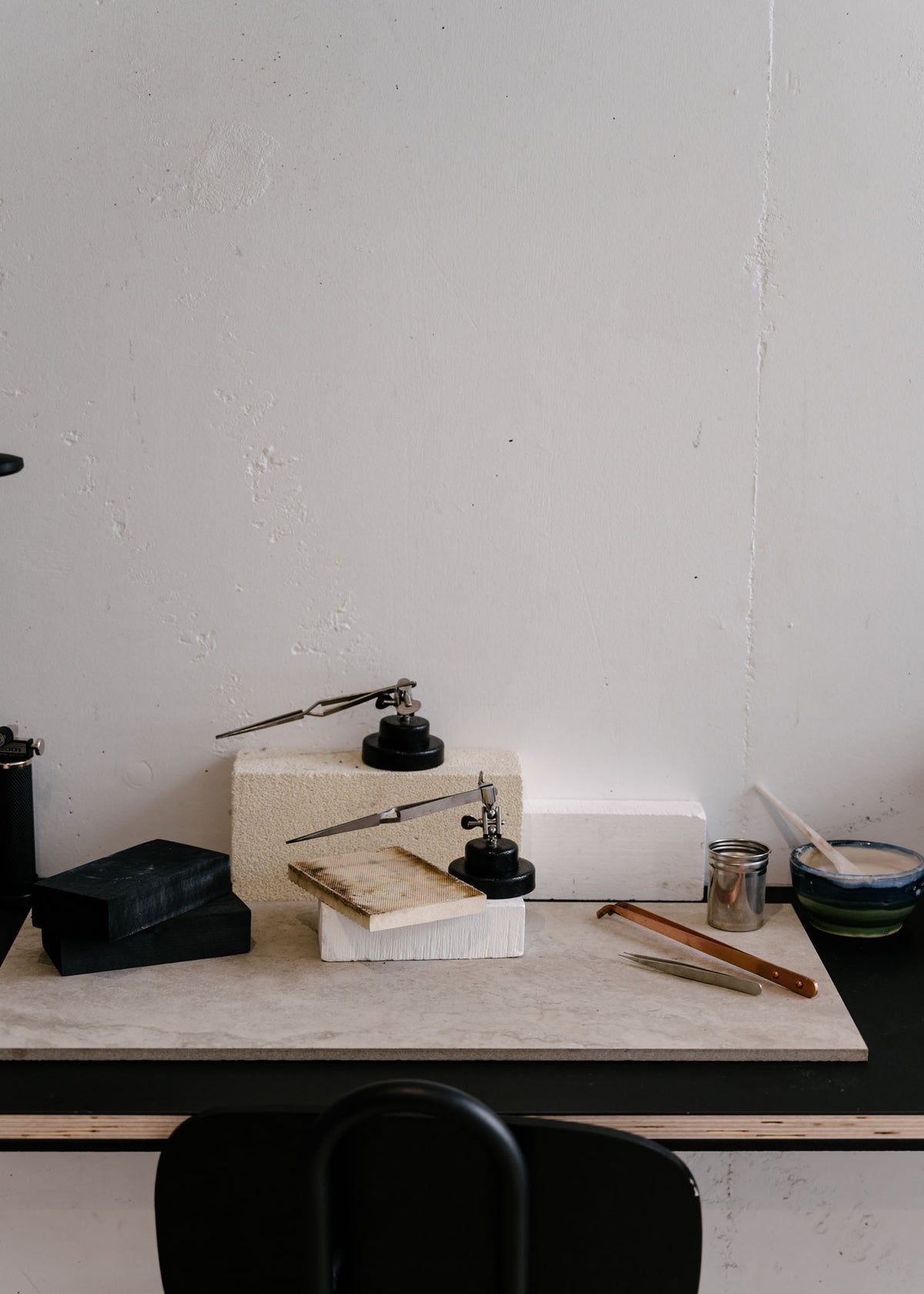 A minimalist jewellery workbench with white bricks, black tools, and small containers placed on a sleek black surface against a plain wall.