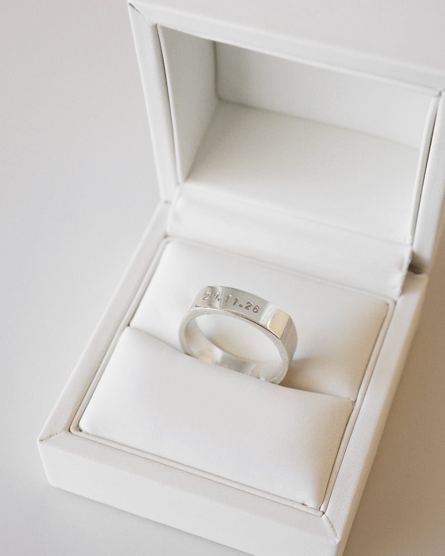 Silver ring with engraved text in an open white jewelry box on a light gray background
