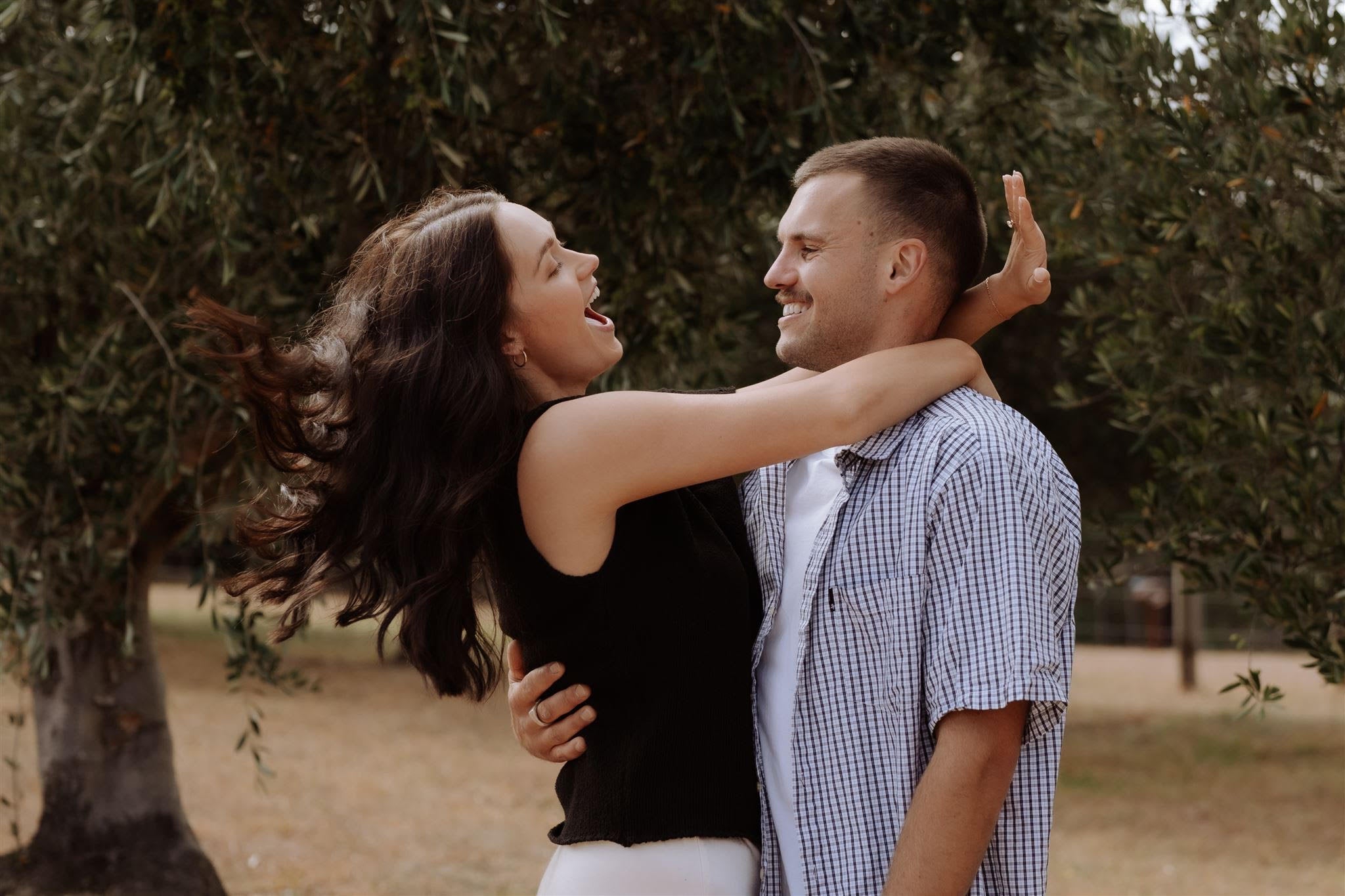 A couple embraces joyfully in a park, the woman showcasing a ring while smiling at her partner, surrounded by lush trees.
